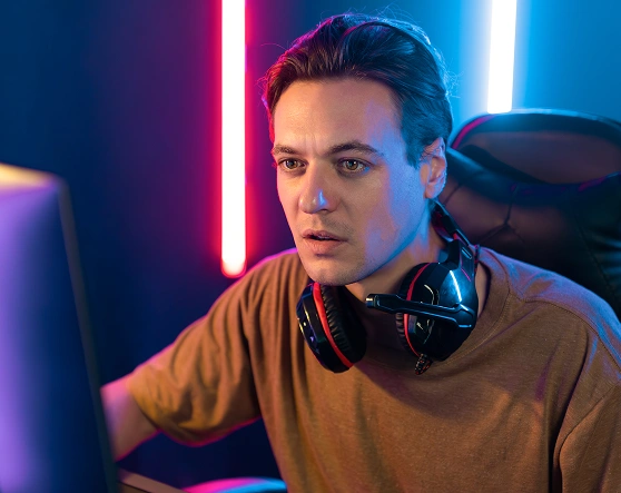 Iemand met een koptelefoon om de nek kijkt geconcentreerd naar een computerscherm. Blauwe en roze neonlichten werpen een levendige gloed in de kamer.