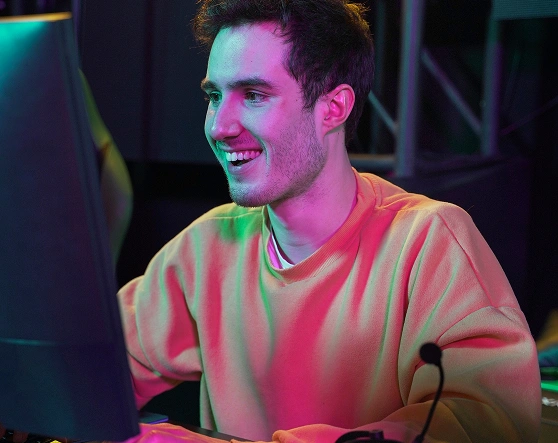 Een jonge man in een gele trui glimlacht terwijl hij typt op een toetsenbord onder felroze en blauw licht. Zijn koptelefoon ligt op tafel en creëert een energieke game-sfeer.