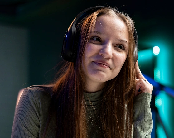 Een persoon met lang haar zit met een hoofdtelefoon op zachtjes te glimlachen in een schemerige kamer met blauwgroene accenten. De sfeer is ontspannen en bezinnend.